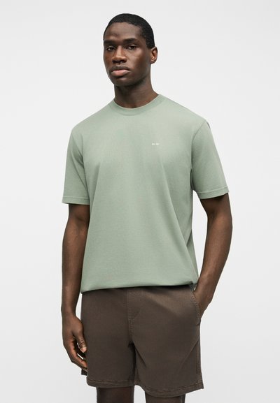 Homme portant un t-shirt vert sauge à manches courtes avec un petit logo blanc et un short marron foncé, debout avec une main dans la poche sur fond blanc.
