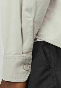 Manche de veste en coton beige avec un poignet à bouton. Présente des détails de couture et une finition lisse, légèrement texturée. Tissu noir visible en dessous.