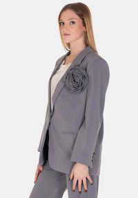 Blazer grigio con una grande rosa in tessuto accentuata sulla spalla sinistra. Taglio sartoriale, maniche lunghe, chiusura con un singolo bottone, con una texture morbida.