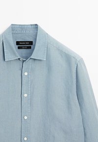 Chemise en lin bleu clair avec un col classique, fermeture à bouton sur le devant et boutons blancs. Présente une texture lisse et un design décontracté.
