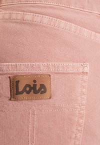 Růžová džínovina s vertikálním žebrováním, s hnědou koženou nášivkou označenou "Lois." Viditelné švy a detaily zadní kapsy.