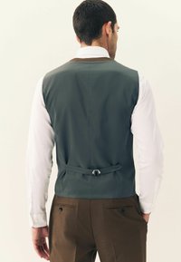 Vest din material gri cu o textură netedă, curea ajustabilă la spate, purtat peste o cămașă albă, asortat cu pantaloni maro care au două buzunare la spate.