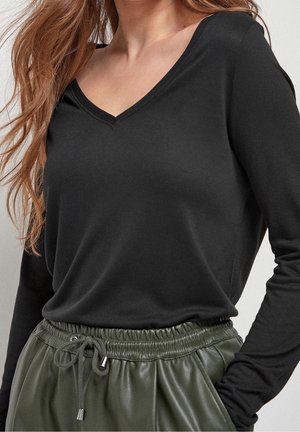 Femme portant un haut à manches longues noir avec col en V, rentré dans un pantalon taille haute vert olive à cordons avec une texture façon cuir.