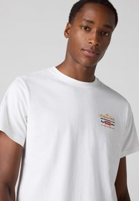 Witte katoenen t-shirt met een multikleurige geborduurde logo op de borst. Korte mouwen en een klassieke ronde hals.