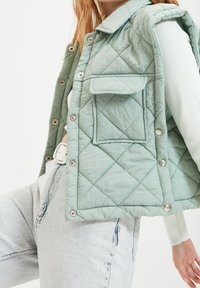 Pikowana miętowo-zielona kamizelka z zatrzaskiem, z jedną przednią kieszenią i zaokrąglonym kołnierzykiem. Nosi się ją na lekkiej bluzce i dżinsowych spodniach.