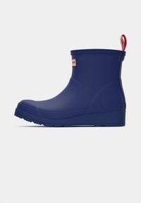 Cizmă de ploaie din cauciuc albastru, cu un finisaj neted, având un patch cu un logo roșu și alb și un tab pentru a fi ușor de purtat la spate. Design scurt.