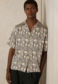 Cămașă cu mânecă scurtă dintr-un material crem, având un model îndrăzneț în valuri de culoare bleumarin, cu guler și închidere cu nasturi, asortată cu pantaloni negri.