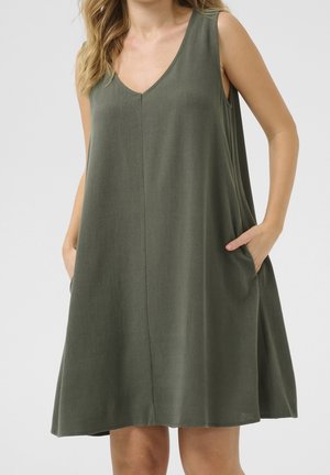 Femme portant une robe olive ample sans manches avec un col en V et les mains dans les poches latérales sur un fond uni.
