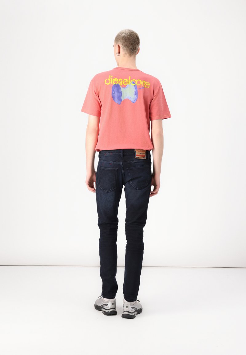 T-shirt en coton rouge avec le logo "dieselcore" et un graphique bleu au dos, associé à un jeans skinny bleu foncé et des baskets grises.