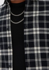Camicia a quadri bianca e nera sopra una maglia nera, con due collane a catena d'argento attorno al collo.