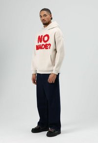 Beige hoodie met rood, pluizige tekst "NO MADE?" op de voorkant, kangoeroezak, comfortabele pasvorm. Gecombineerd met donkeren denim broek met wijde pijpen en zwarte schoenen.