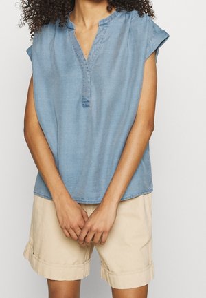 Vrouw draagt een lichtblauw shirt met korte mouwen en beige shorts met opgerolde zoom, staand met handen losjes voor zich gevouwen.