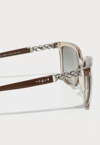 Ochelari de soare cu rame transparente și accente maronii. Prezintă un design cu lanțuri din metal pe brațe și logo-ul "vogue" gravat pe unul dintre brațe.