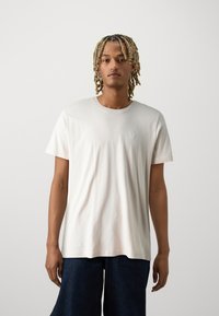 Camiseta de algodón color beige claro con cuello redondo y mangas cortas, que presenta un pequeño logo en el pecho y un corte relajado.