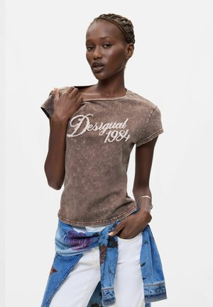 Kobieta z zaplecionymi włosami, ubrana w brązowy t-shirt "Desigual 1984" i białe spodnie, z niebieską kwiecistą kurtką zawiązaną wokół talii.