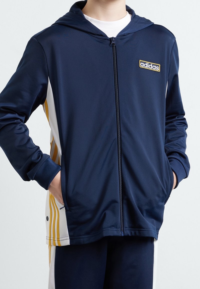 Sudadera con cremallera frontal en color navy, con un panel lateral blanco y rayas amarillas, y un logo bordado en el pecho. Tejido suave, fit regular.