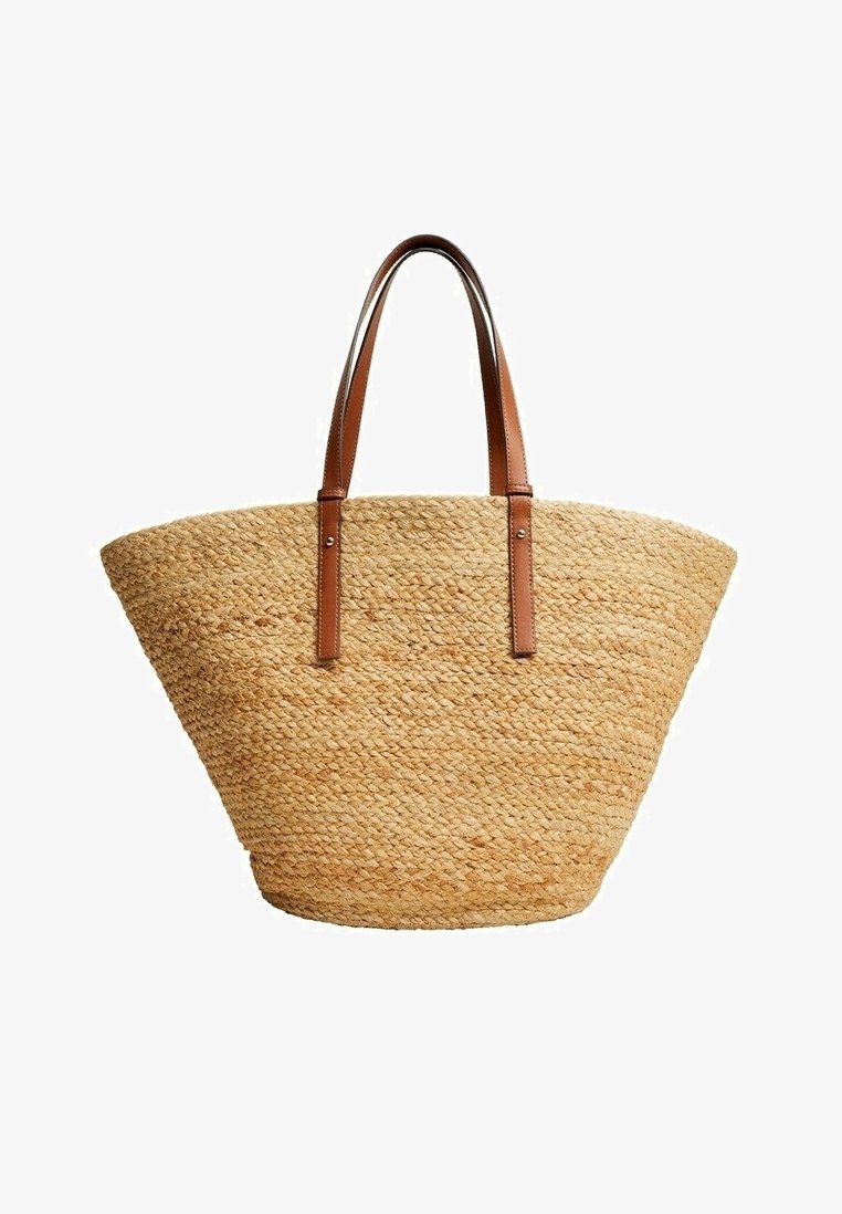 Woven straw tote bag o szerokim, trapezoidalnym kształcie z dwiema brązowymi skórzanymi uchwytami przymocowanymi metalowymi nitami.