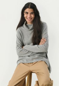 Pulover gri cu guler tip cowl, realizat dintr-un material moale, cu mâneci lungi și croi lejer. Purtați cu pantaloni bej, așezat pe un scaun de lemn.