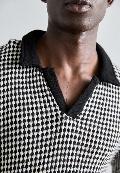 Polo de patrón en blanco y negro con un diseño texturizado, que presenta un collar negro y un escote en forma de V. Primer plano del escote de la prenda.