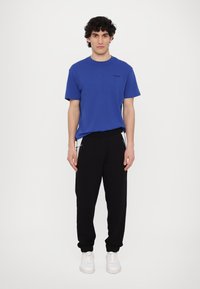 Μπλε κοντομάνικο μπλουζάκι με στρογγυλή λαιμόκοψη, συνδυασμένο με μαύρες φόρμες που διαθέτουν φερμουάρ και ελαστικούς μανσέτες. Τα αθλητικά παπούτσια ολοκληρώνουν το σύνολο.