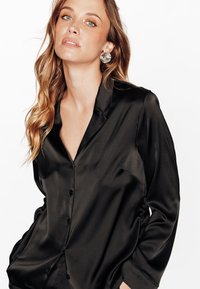 Chemise noire en satin à boutons avec col, poche poitrine unique et manches longues, présentant une texture lisse et une coupe ajustée.