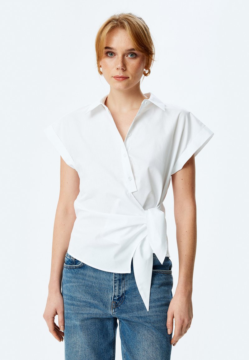 Camicia bianca a maniche corte con colletto, presenta un dettaglio a cravatta in vita. Realizzata in tessuto morbido, ha una vestibilità rilassata e un design minimale.