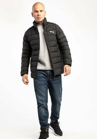 Czarna puchowa kurtka z zapięciem na zamek i poziomym pikowaniem, noszona na szarym swetrze i niebieskich dżinsach; posiada logo Puma.