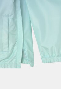 Light turquoise windbreaker with a smooth, shiny texture. Features elastic cuffs, a zippered pocket, and a mesh lining.
