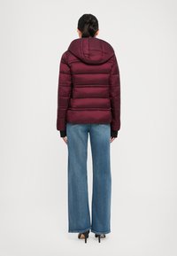 Woman wearing a maroon hooded puffer jacket, blue wide-leg jeans, and black high-heeled shoes, standing facing away against a white background.