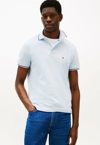 Polo azul claro com colarinho, apresentando detalhes riscas em azul-marinho e branco, mangas curtas, e um pequeno logótipo no peito.