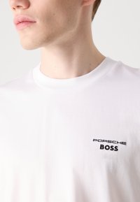 Biela bavlnená tričko s okrúhlym výstrihom, s čiernym vyšitou značkou "PORSCHE" a "BOSS" na ľavej strane hrude. Mäkká textúra, voľný strih.