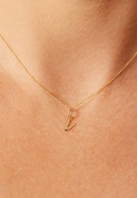 Gouden ketting met een gestileerde hanger in de vorm van de letter "V". De ketting is dun met een gladde textuur en de hanger is glanzend en geometrisch.