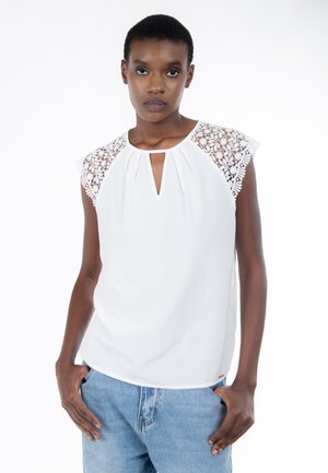 Donna che indossa una maglia bianca senza maniche con spalle in pizzo e jeans blu, in piedi con le mani nelle tasche davanti a uno sfondo bianco uniforme.