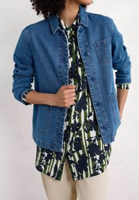Veste en denim à coupe décontractée, de couleur bleue, avec des boutons à l'avant et deux poches latérales, portée sur une chemise à motifs floraux verts et marins.