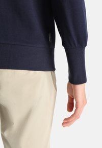 Navy blue sweater featuring ribbed cuffs and hem, with a soft texture. Beigecolored pants partially visible, hands positioned around fabric edges.