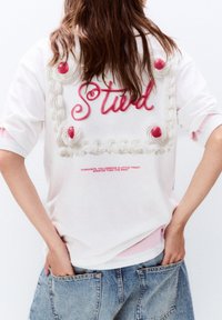 Hvid kortærmet t-shirt med et design af pink glasur med ordet "Stud" og kirsebær, fremhævet med yderligere tekst nedenfor.