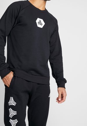 Uomo in camicia a maniche lunghe nera e pantaloni con loghi geometrici bianchi, mano in tasca, su uno sfondo grigio chiaro uniforme.