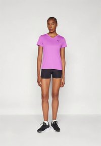 En kvinne står iført en lilla V-hals T-skjorte, svarte shorts, hvite sokker og svarte joggesko mot en enkel lys bakgrunn.