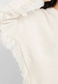 Sweter w kolorze kremowym, dziany, z frędzlami na rękawach i ribbowym wykończeniem. Miękka tekstura, casualowy, luźny krój.