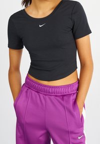 Czarny krótki rękaw crop top z okrągłym dekoltem, z małym białym logo Nike na klatce piersiowej, w połączeniu z fioletowymi spodniami dresowymi.