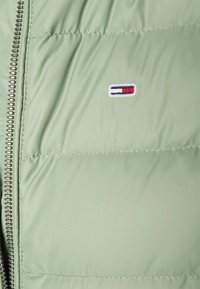 Zielona puchowa kurtka z zamkiem błyskawicznym i poziomym pikowaniem. Tkanina jest gładka, z małym haftowanym logo w kolorach czerwonym, białym i niebieskim.