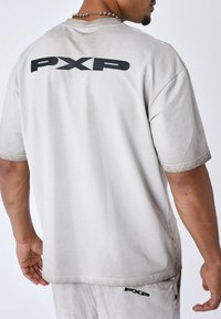 Camiseta de corte holgado en gris claro, confeccionada en algodón, con el logo negro "PXP" en la parte posterior. Diseño de mangas cortas y cuello redondo. Sin patrones destacados.