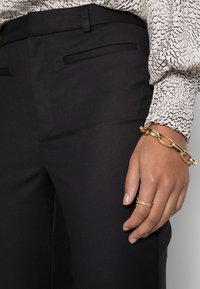 Czarne dopasowane spodnie z bokami, gładka faktura i przyległy krój. Na boku znajduje się ramię z złotymi akcentami łańcucha i pierścionkami.