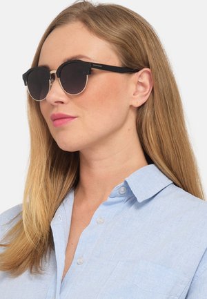 Sonnenbrille mit schwarzen Rahmen und dunklen Gläsern, im runden Design und mit markanter Metallbrücke, kombiniert mit einem hellblauen Hemd mit Kragen.
