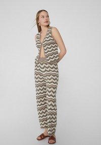 Gebreide vest en broek set met een Chevronpatroon in aardetinten. Het vest heeft een open voorkant; de broek is wijd en heeft een ontspannen pasvorm.