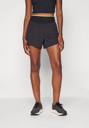 Person, die schwarze Sportshorts, ein schwarzes Crop-Top und schwarze Laufschuhe trägt, steht vor einem einfarbigen hellen Hintergrund.