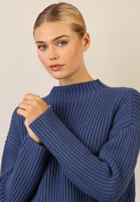 Sweter na drutach w głębokim niebieskim kolorze, z żeberkową teksturą, wysoką szyją i długimi rękawami. Ręce złączone na szyi, podkreślające design.