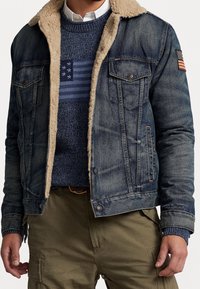 Jeansjacka med fuskpälskrage, mörkblå tvätt och två bröstfickor. Under den, en marinblå stickad tröja med randigt mönster och amerikansk flagga.