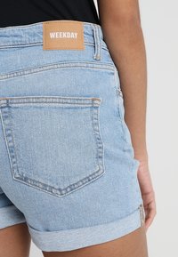 Pantalones cortos de mezclilla azul claro con dobladillos enrollados, que cuentan con una etiqueta de cuero marrón que dice "WEEKDAY" en la parte trasera y bolsillos traseros estándar.