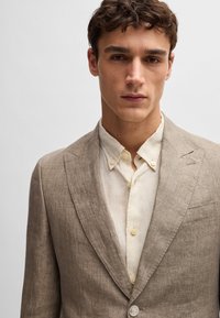 Brown linen blazer with notch lapels, two-button closure, and a pocket. Paired with a light beige button-down shirt.